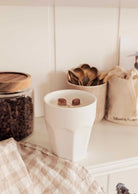 A white cup with milk and two brown cubes sits on a counter near a jar of coffee beans, wooden utensils, a cloth napkin, and the Mimi & August Special Edition Candle, adding warmth to the cozy scene.