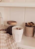 A white cup with a drink and marshmallows sits next to coffee beans, gold spoons, and the Mimi & August Special Edition Candle. A beige checkered cloth nearby adds a cozy charm to the scene.