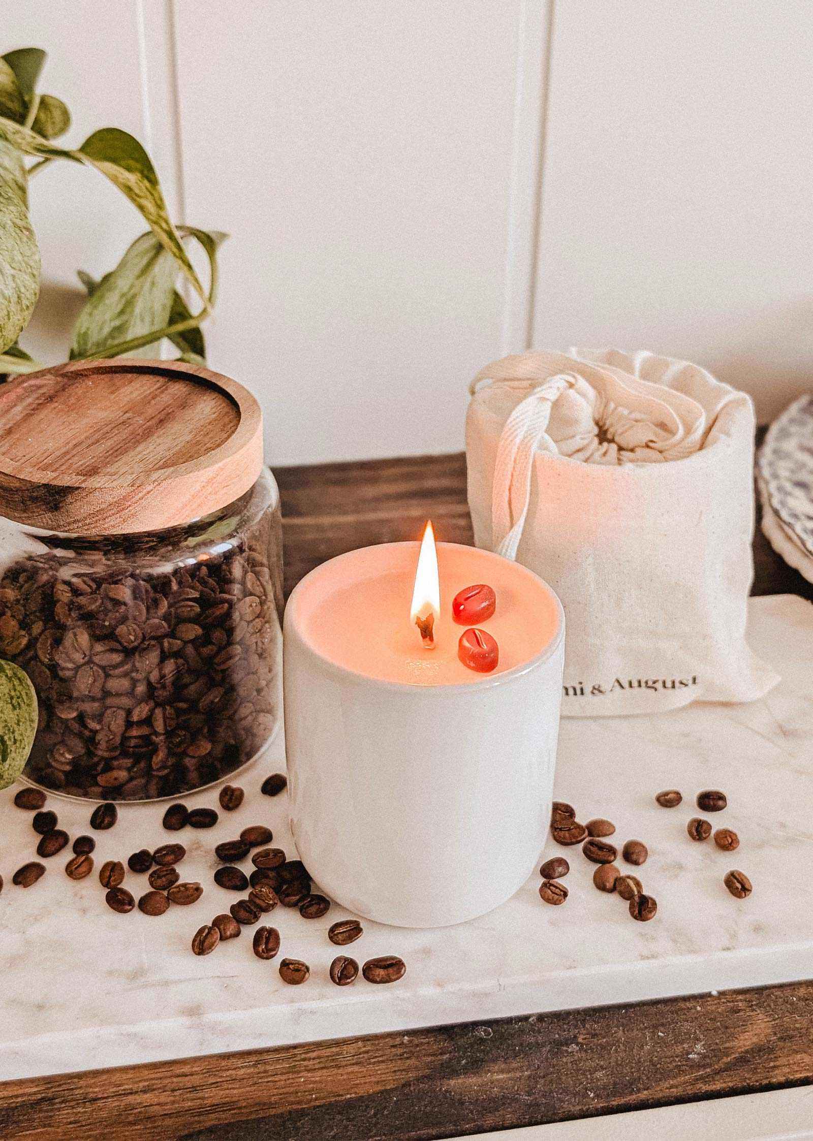 The Special Edition Candle by Mimi & August, adorned with red stones, is lit on a marble tray next to a glass jar and pouch of coffee beans, with loose beans scattered nearby.