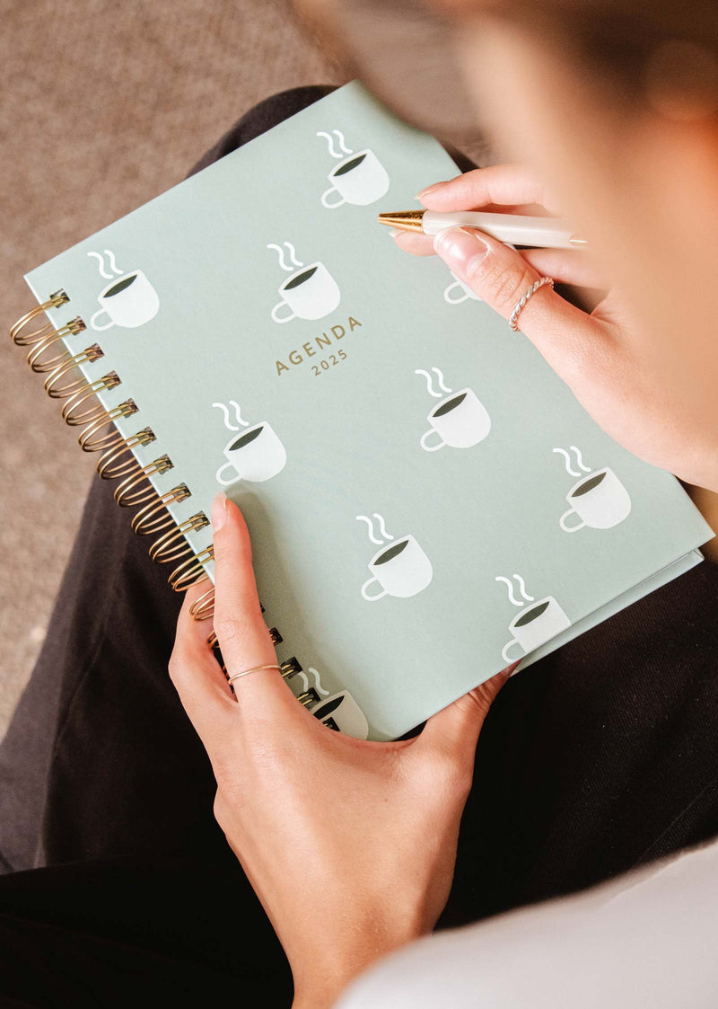 A person holds and writes in the Cafecito - Agenda 2025 planner by Mimi & August, decorated with charming cup illustrations, ideal for coffee enthusiasts.