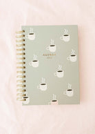 The Cafecito Agenda 2025 from Mimi & August, ideal for coffee enthusiasts, showcases a spiral binding and a delightful coffee cup illustration set against a light green cover, beautifully contrasted with a light pink background.