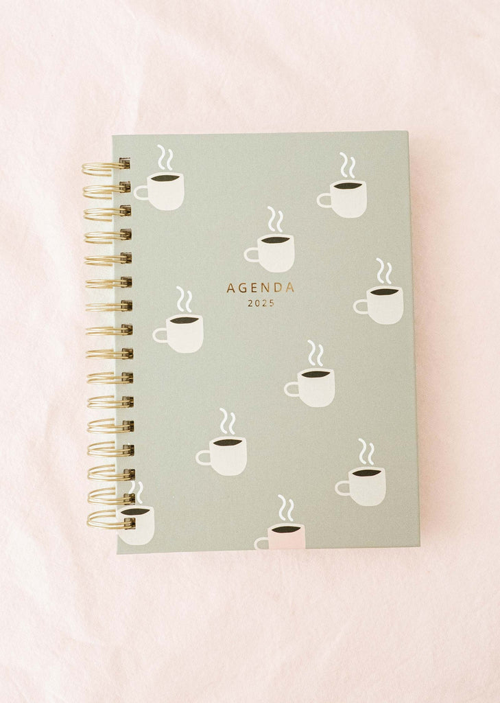 The Cafecito Agenda 2025 from Mimi & August, ideal for coffee enthusiasts, showcases a spiral binding and a delightful coffee cup illustration set against a light green cover, beautifully contrasted with a light pink background.