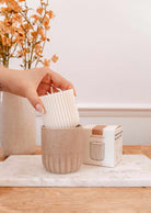 A hand places a Mimi & August Candle Refill Cafecito into a beige ceramic holder beside its box on a table, with a vase of orange flowers in the background.