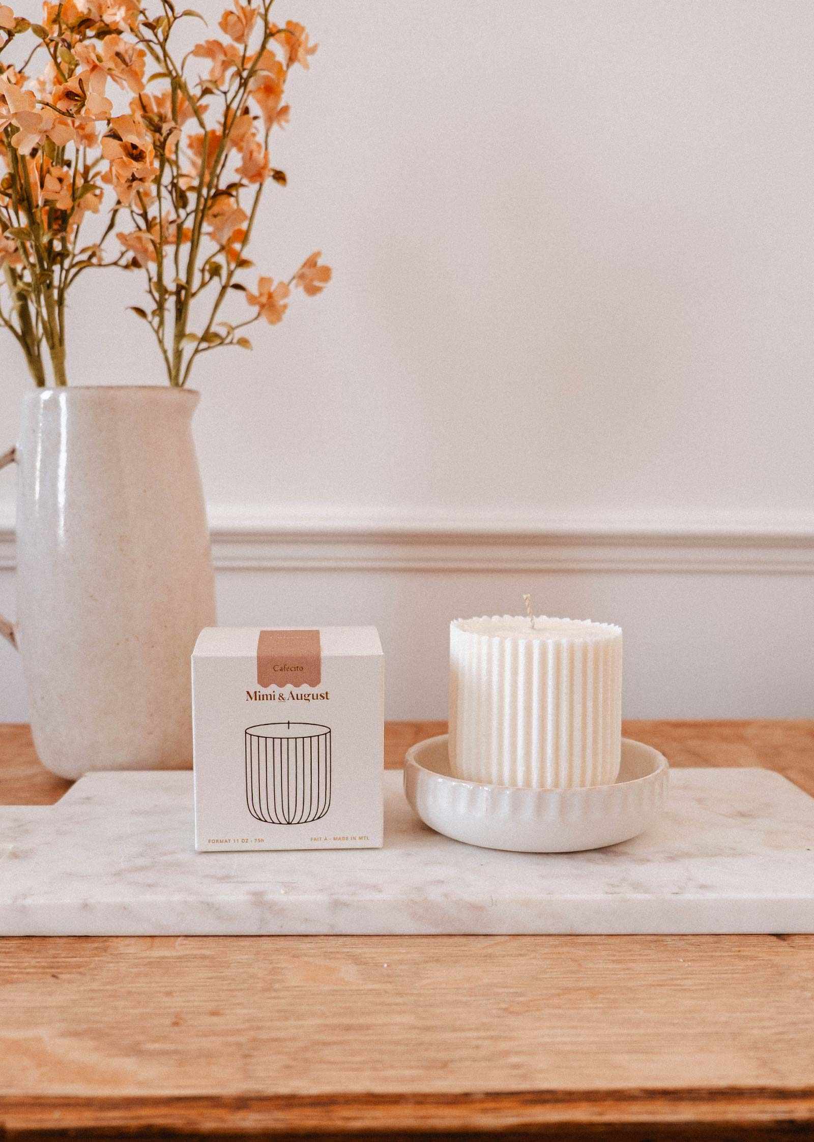 A white ribbed Mimi & August Candle Refill Cafecito, a coffee-scented candle in ceramic, sits beside its box on a marble tray; orange flowers in a vase are visible on the wooden table in the background.