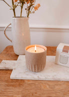 The Candle Refill Cafecito by Mimi & August, in a beige ceramic holder, is lit on a marble board with flowers and a white box in the background atop a wooden surface.