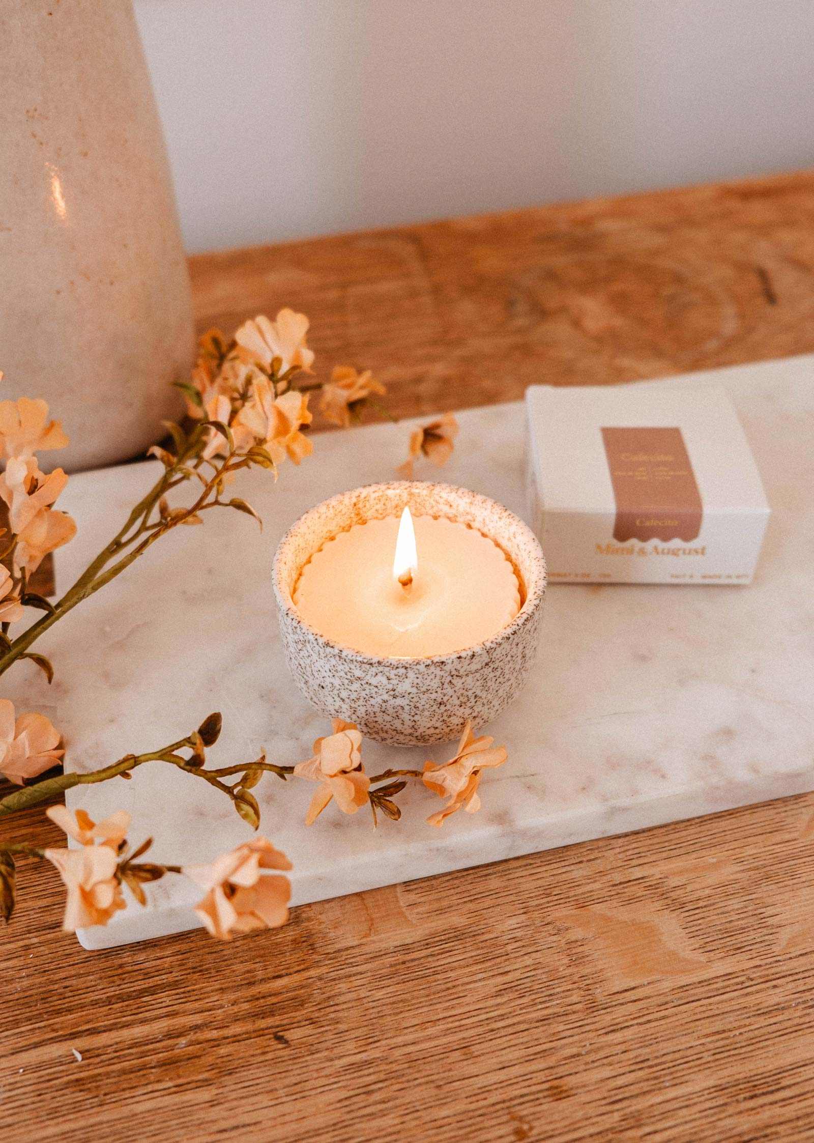 A lit Mimi & August Candle Refill Cafecito in a textured holder sits on a marble tray beside peach flowers and a box, arranged on a wooden surface.