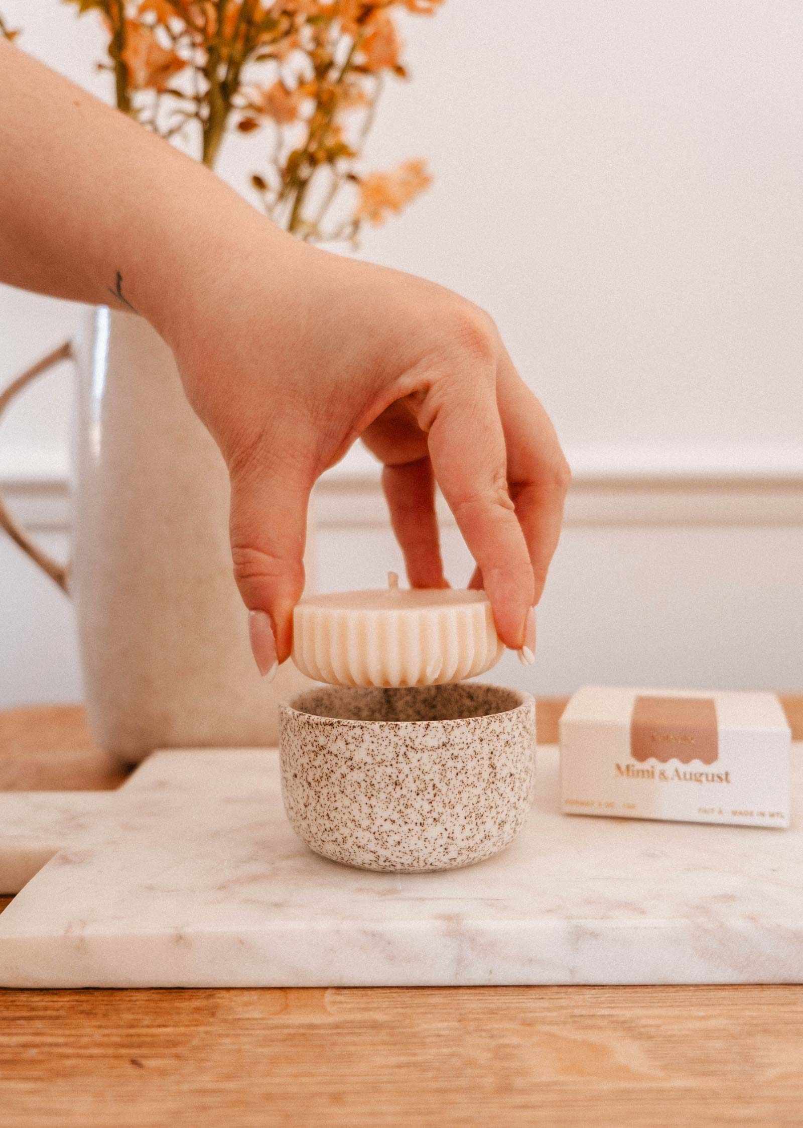 A hand lifts a ribbed white Candle Refill Cafecito by Mimi & August from a speckled ceramic holder on a marble tray, with a flower vase and a Mini August candle box in the background.