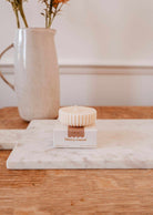 A ribbed cream-colored Candle Refill Cafecito by Mimi & August sits on a white box, placed on a marble board with a ceramic vase and flowers in the background.
