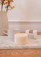 The Candle Refill Cafecito by Mimi & August, a ribbed cream-colored soy wax candle, rests on a marble slab beside a ceramic mug, a Musk Accord box, and a vase of orange flowers.