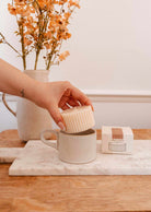 A hand places a ribbed Mimi & August Candle Refill Cafecito into a beige mug on a marble board, with its box and a vase of orange flowers in the background.