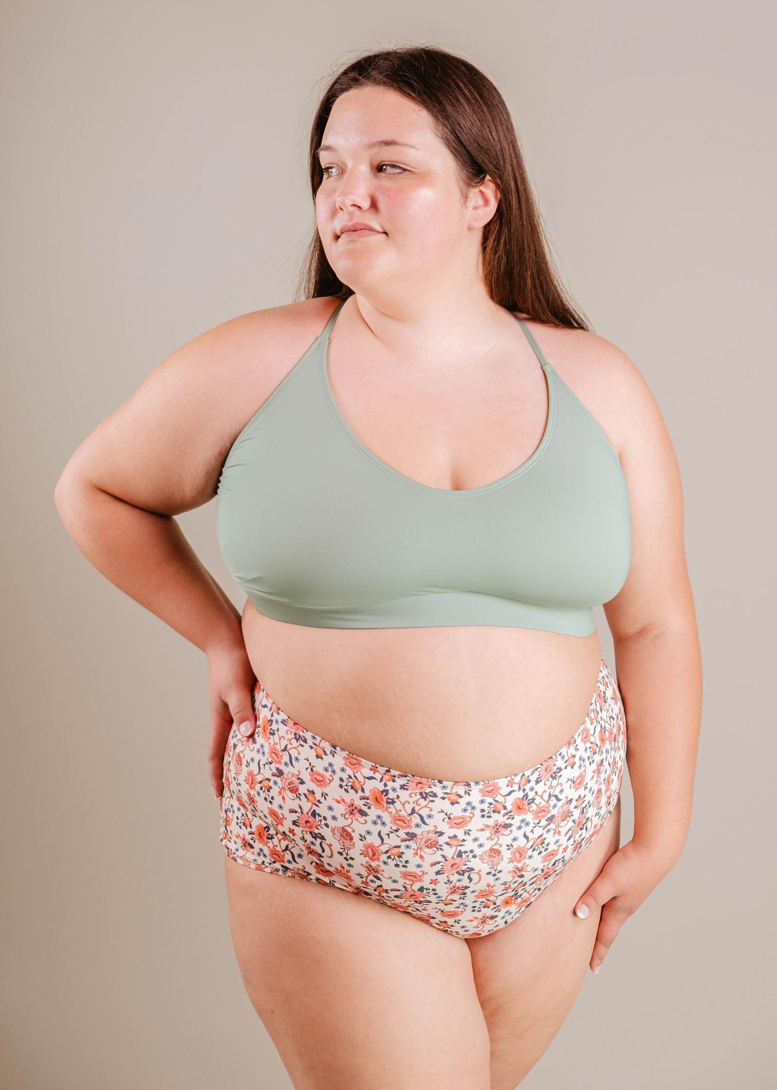 A young woman in a green Mimi & August Chichi Agave Bralette Bikini Top and floral print bikini bottoms stands confidently, looking to the side against a neutral backdrop.