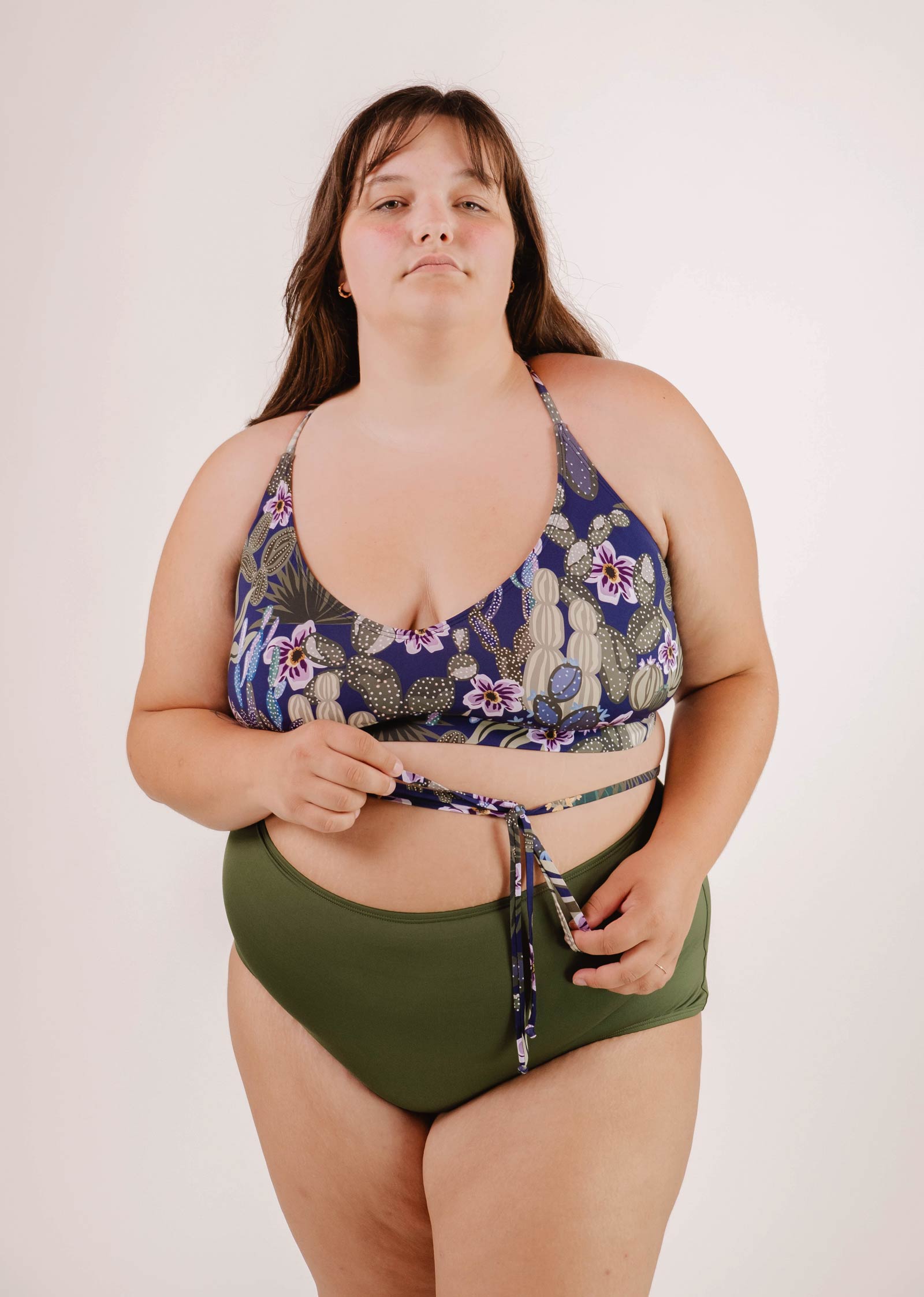 A woman in a green and floral tropical print Mimi & August Chichi Jardin de Nuit - Bralette Bikini Top stands against a plain background, holding the strap of her bikini top as she looks at the camera.