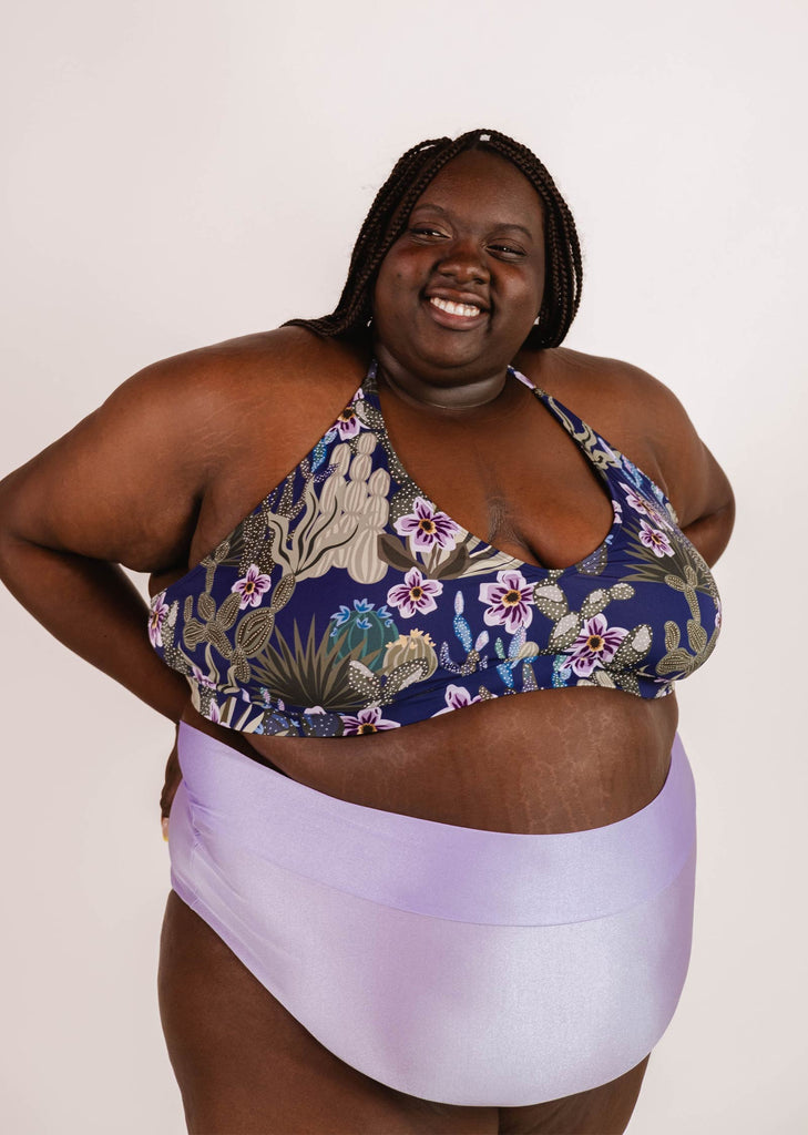 A person with braided hair smiles while wearing a Chichi Jardin de Nuit - Bralette Bikini Top by Mimi & August and light purple bottoms against a plain background.