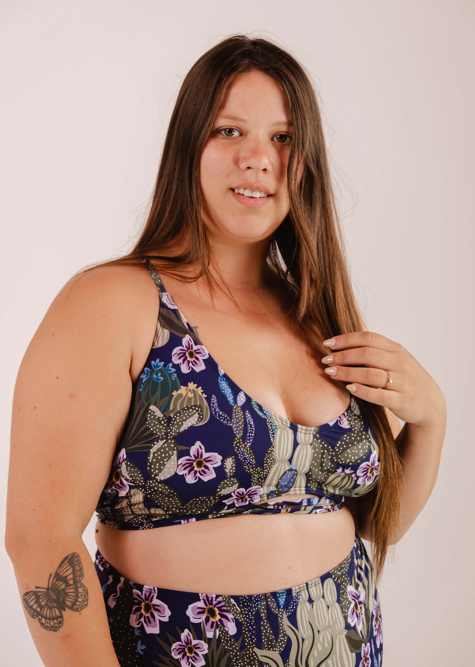 A woman with long brown hair is wearing a Mimi & August Chichi Jardin de Nuit - Bralette Bikini Top with adjustable straps. She has a butterfly tattoo on her arm and is standing against a plain background, looking at the camera.