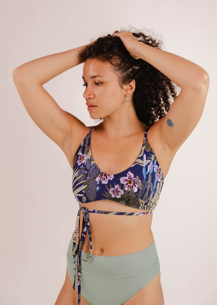 A woman in a Chichi Jardin de Nuit - Bralette Bikini Top by Mimi & August with adjustable straps and green bottoms poses with her hands on her head against a plain background. She has a small bandage on her left arm.