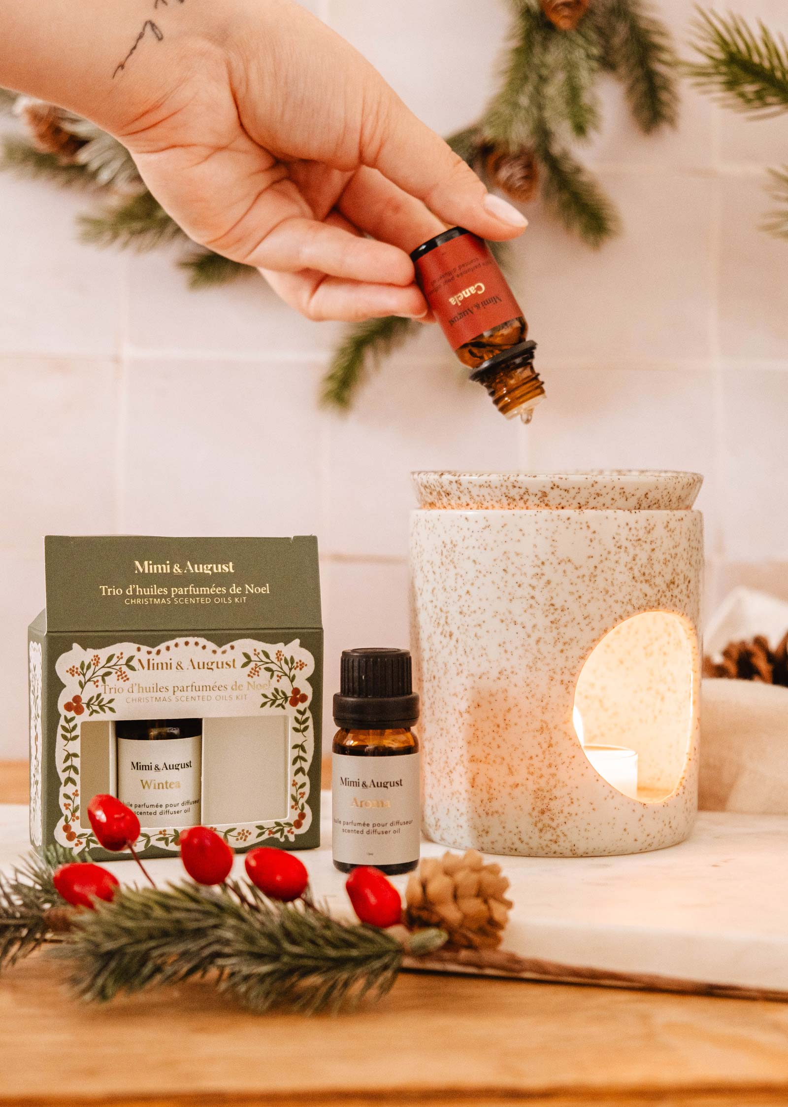 A hand pours essential oil into a white ceramic diffuser, while the *Set | Christmas Scented Oils Trio* by Mimi & August box and two oil bottles are on the table amidst pine needles and red berries.