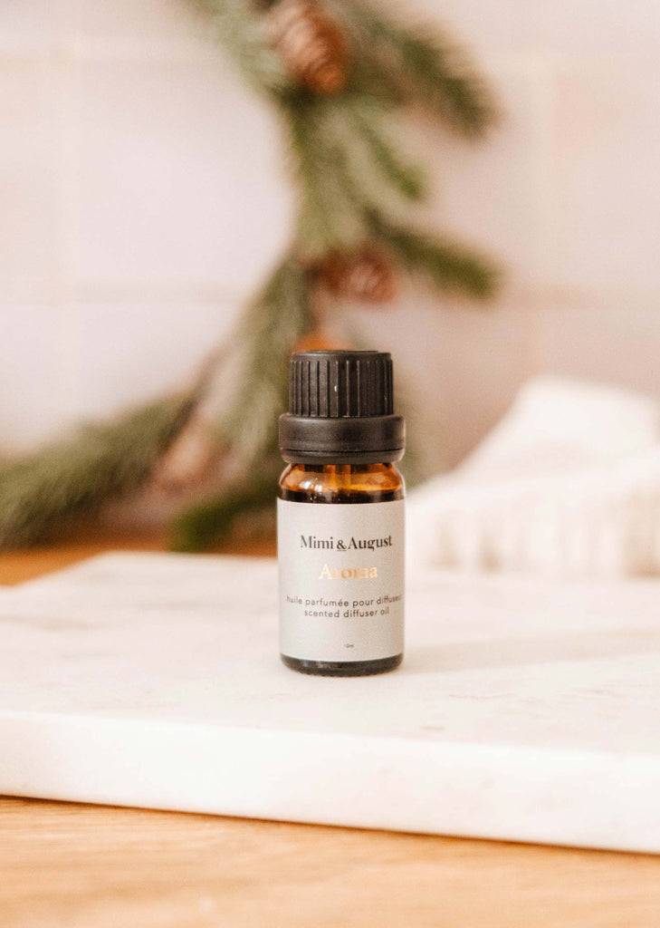 A small bottle from the Christmas Scented Oils Trio by Mimi & August on a white surface, with a blurred background of pine branches.