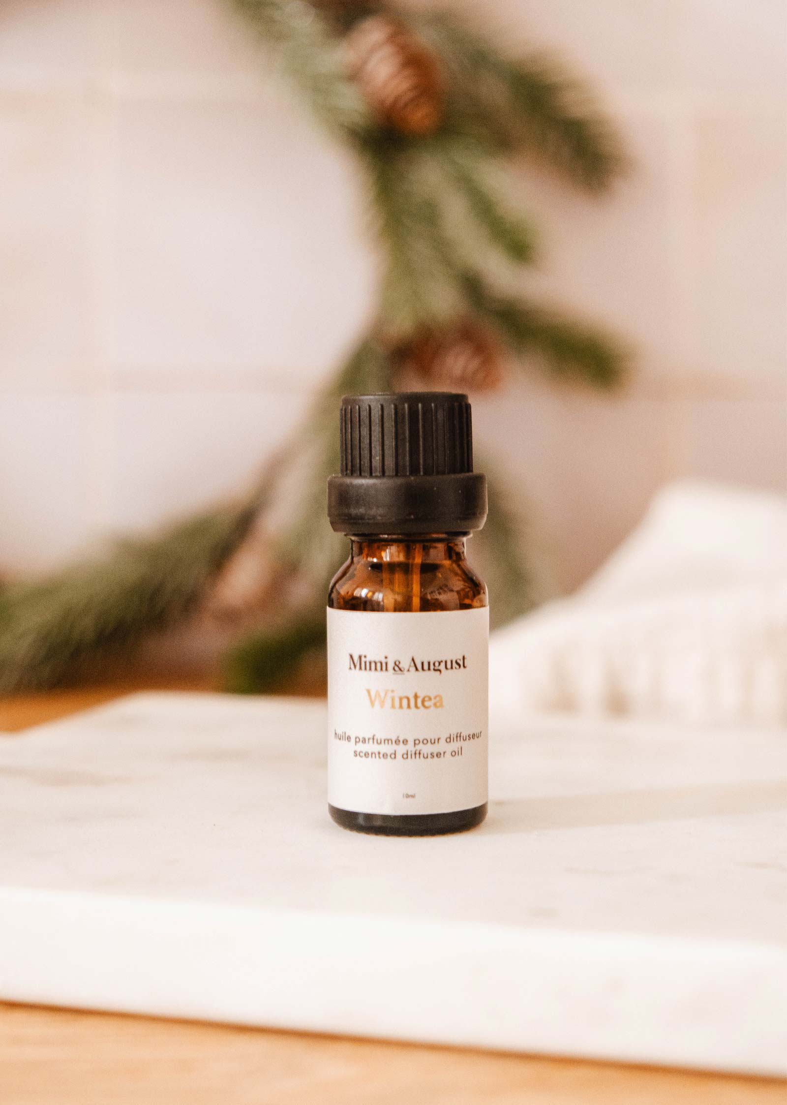 A small amber bottle from the Christmas Scented Oils Trio by Mimi & August, placed on a light surface, with some blurred greenery in the background.