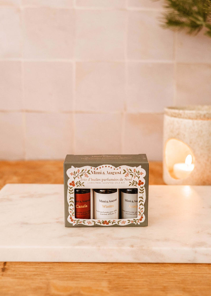 A gift set of the Christmas Scented Oils Trio by Mimi & August, elegantly packaged in a decorative box, rests on a marble surface with a lit candle softly glowing in the background.