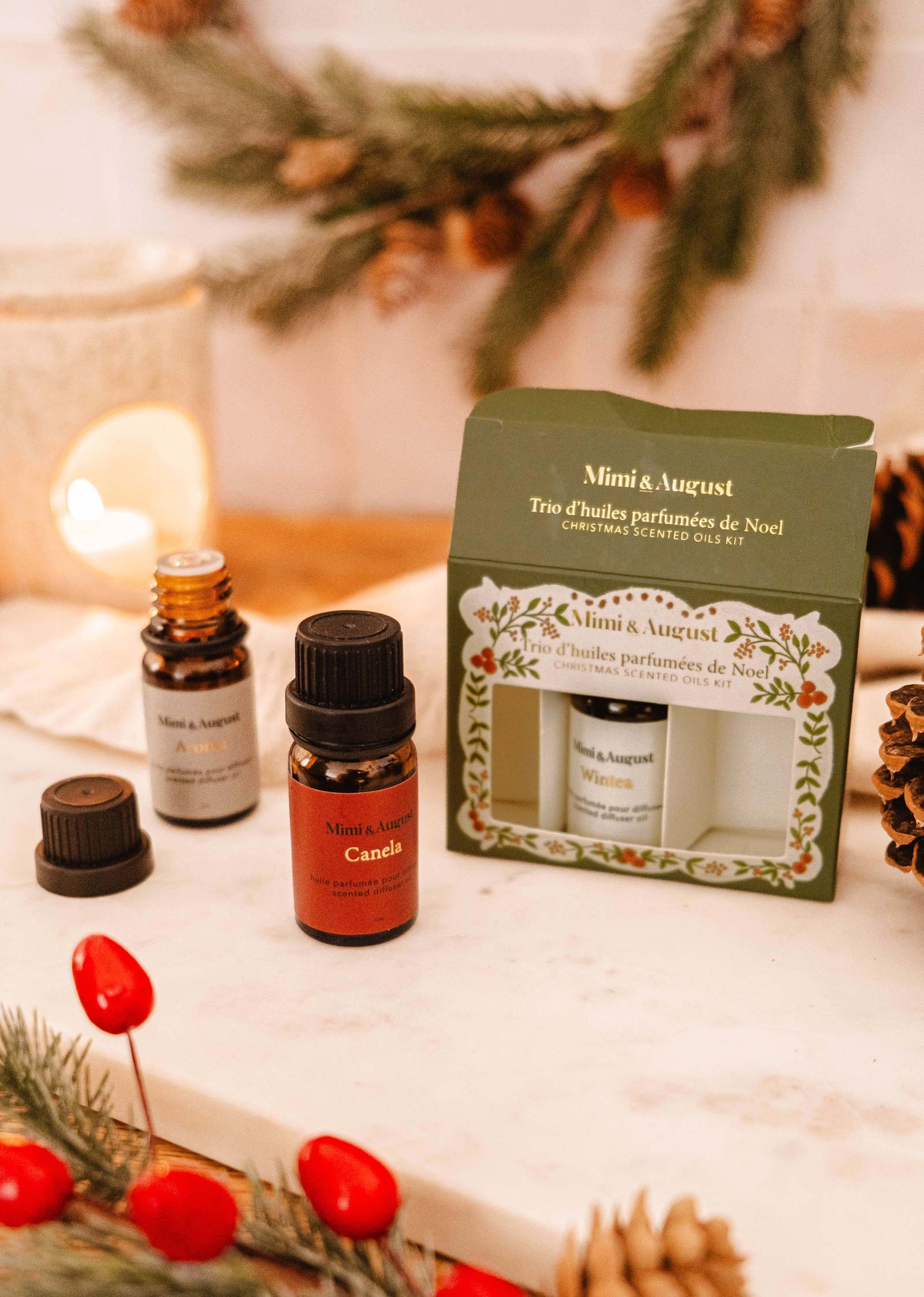 A set of Christmas Scented Oils Trio by Mimi & August is showcased on a table adorned with festive decorations, featuring greenery and red berries.