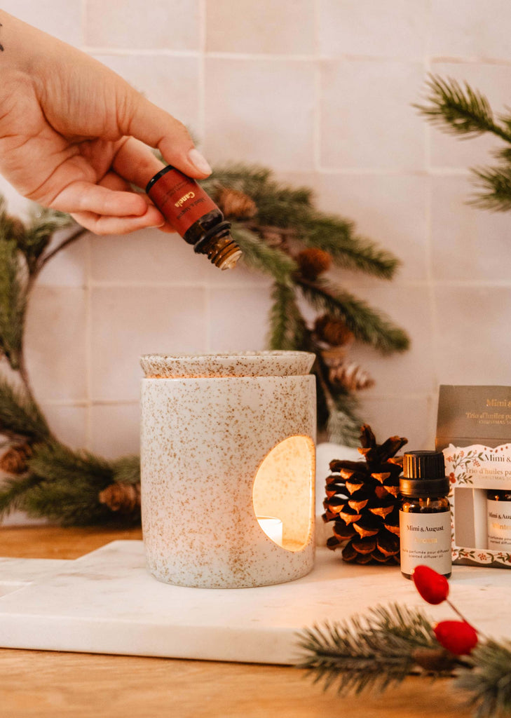 A hand delicately pours Christmas Scented Oil from the Mimi & August trio set into a ceramic diffuser, while holiday greenery and pinecones add festive charm to the background.