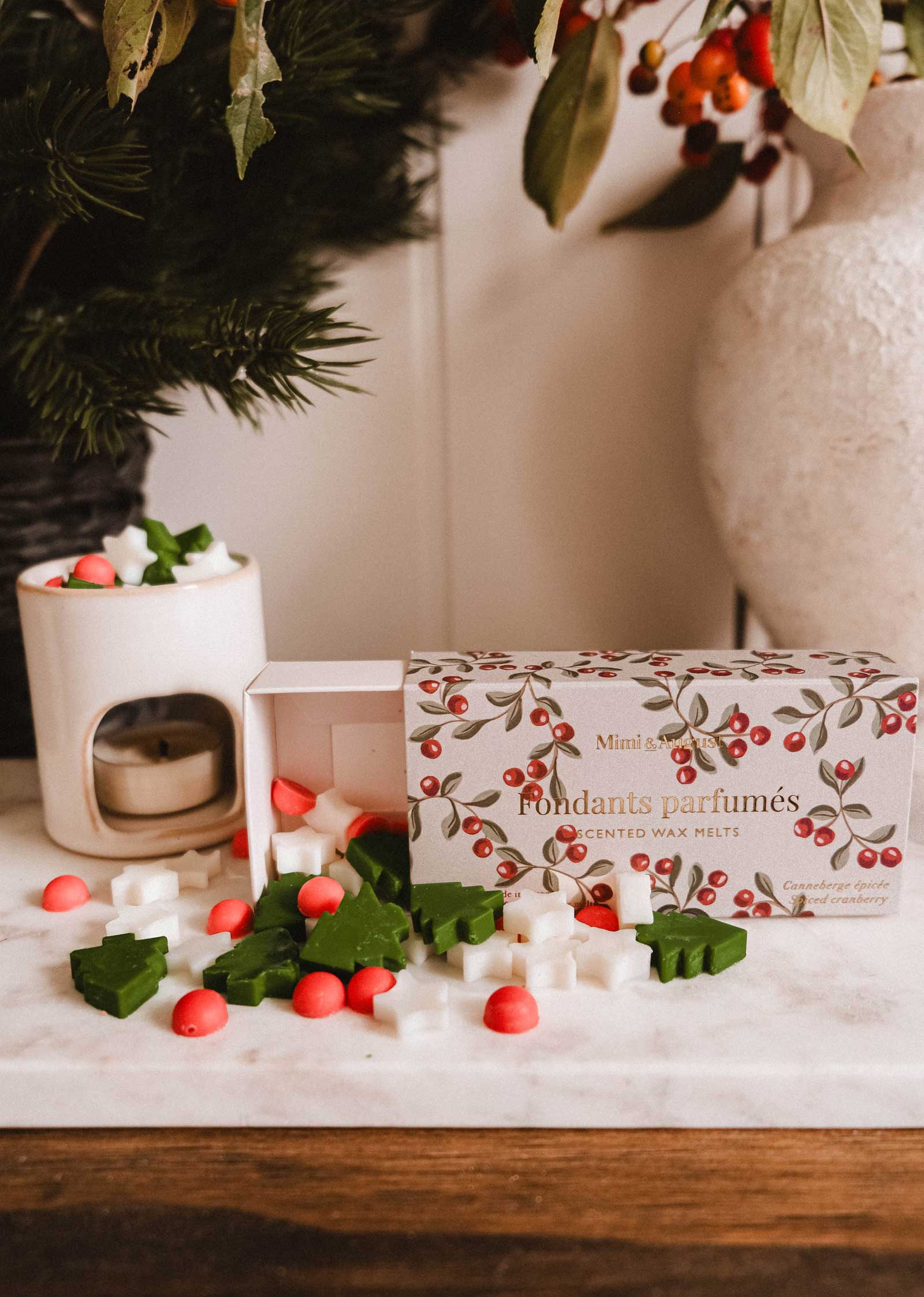 An open box of Mimi & August’s Christmas Scented Wax Melts—shaped like stars, leaves, and berries—sits on a marble surface next to a wax warmer and decorative plants.