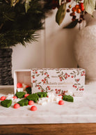 A box of Mimi & August Christmas Scented Wax Melts rests on a marble surface, with green, white, and red holiday wax melts—including spiced cranberry—scattered in front.