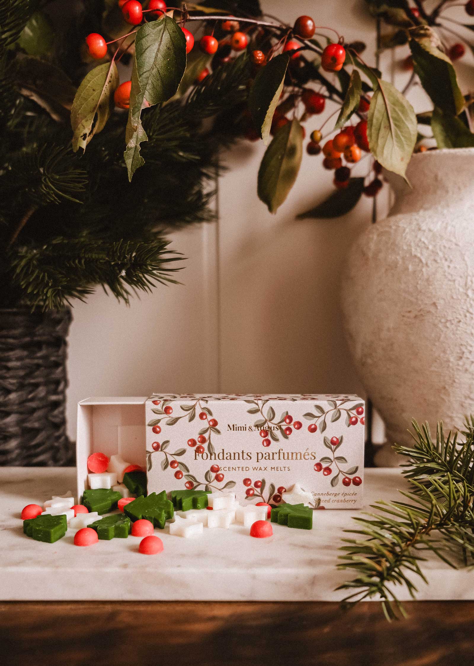 Christmas Scented Wax Melts by Mimi & August, in green, white, and red star and circle shapes, are displayed on a wooden surface beside pine branches and a ceramic vase with berry stems.
