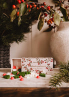 Christmas Scented Wax Melts by Mimi & August, in green, white, and red star and circle shapes, are displayed on a wooden surface beside pine branches and a ceramic vase with berry stems.