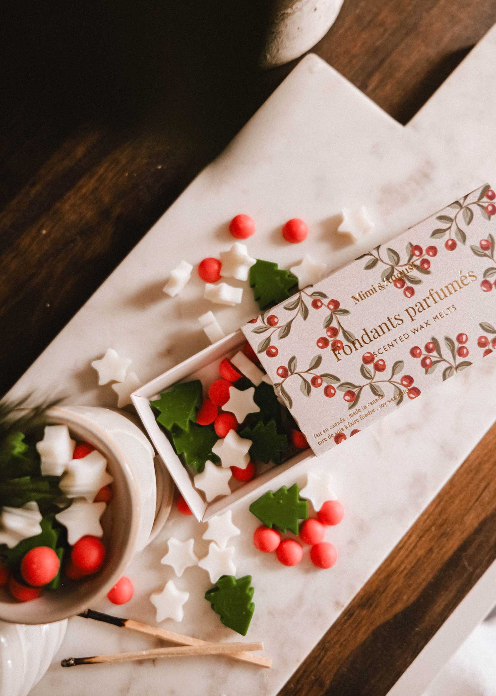 Mimi & August Christmas Scented Wax Melts in festive shapes (stars, trees, red circles) are displayed on a marble tray alongside an open decorative box and matches.