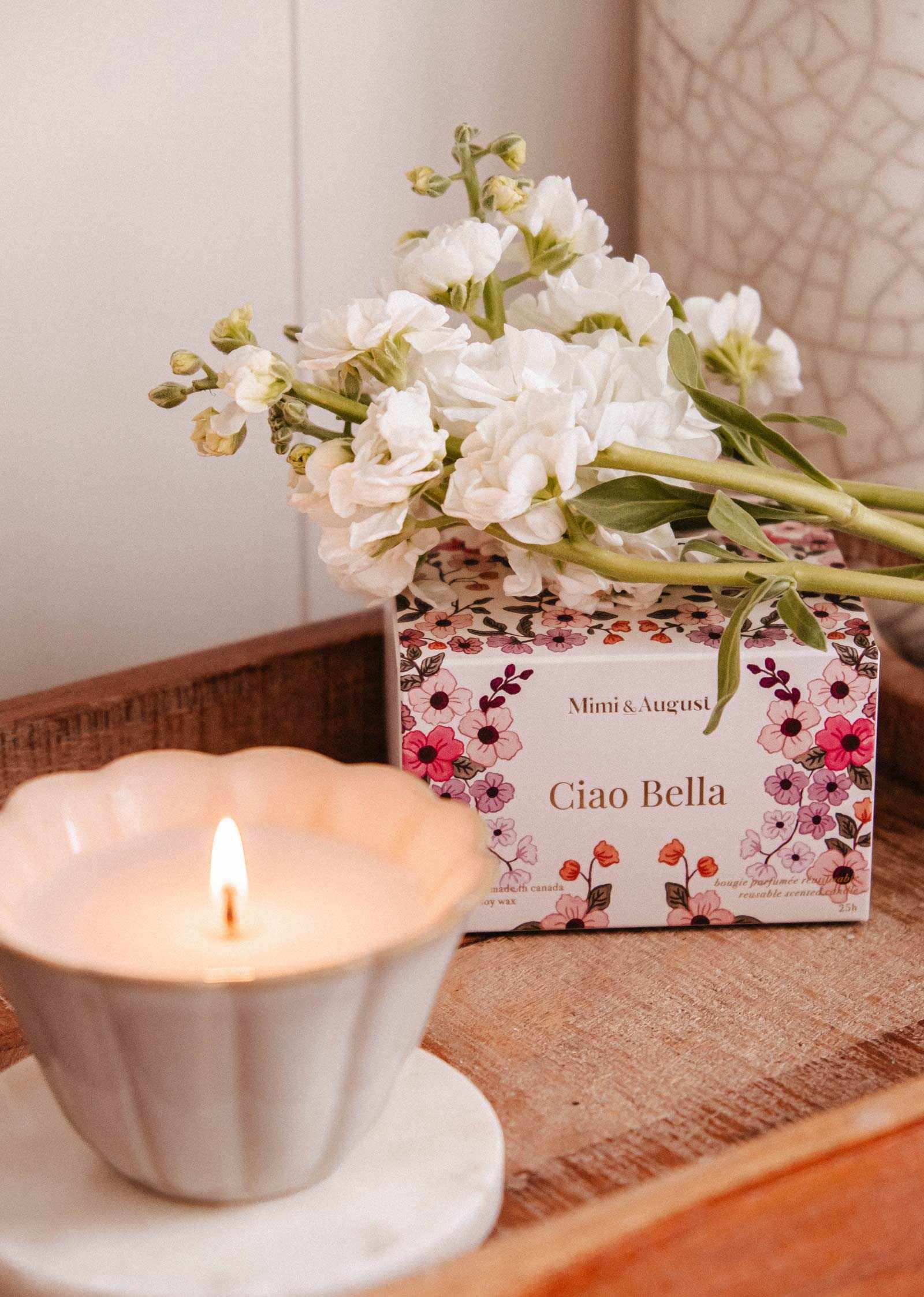 A Mimi & August Ciao Bella Reusable Candle in a ceramic jar sits on a wooden tray next to a floral "Ciao Bella" box, topped with white flowers.
