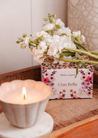 A Mimi & August Ciao Bella Reusable Candle in a ceramic jar sits on a wooden tray next to a floral "Ciao Bella" box, topped with white flowers.