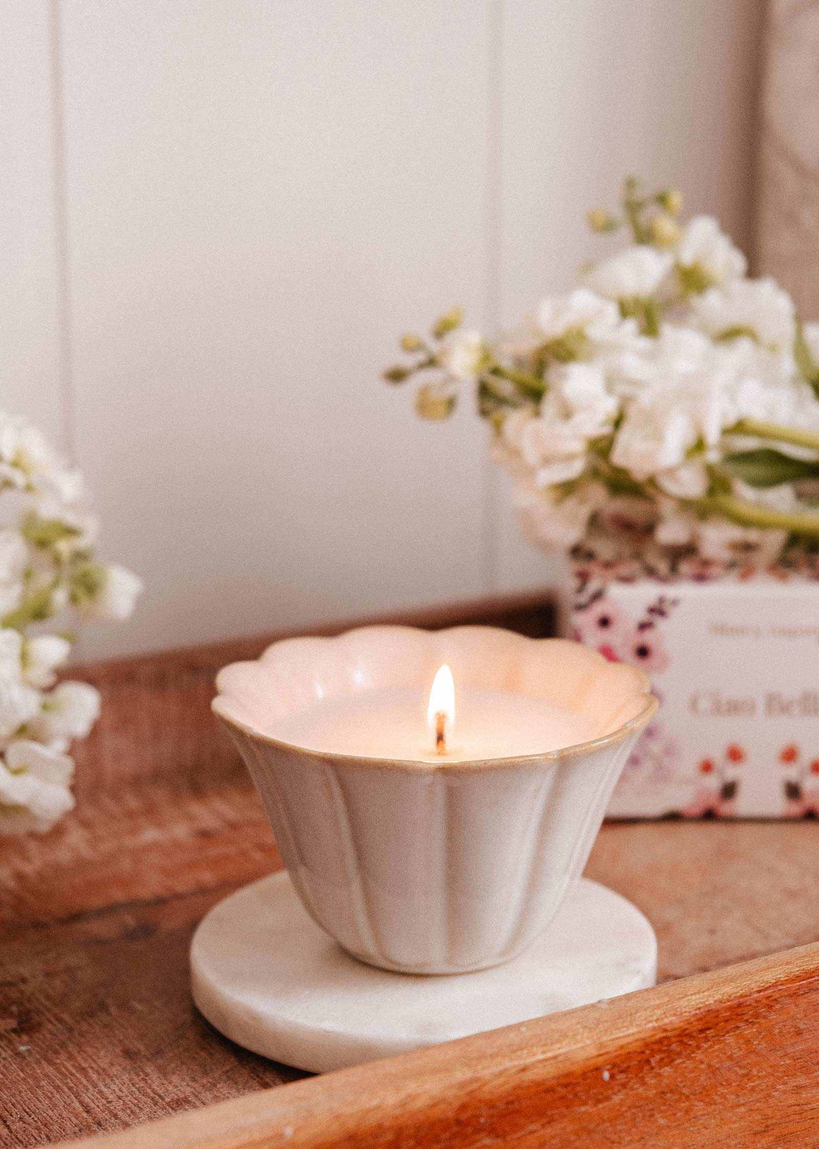 The Ciao Bella - Reusable Candle by Mimi & August, in a white scalloped ceramic cup on a round marble coaster, is displayed with white flowers and a floral box in the background on a wooden tray.