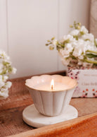 The Ciao Bella - Reusable Candle by Mimi & August, in a white scalloped ceramic cup on a round marble coaster, is displayed with white flowers and a floral box in the background on a wooden tray.