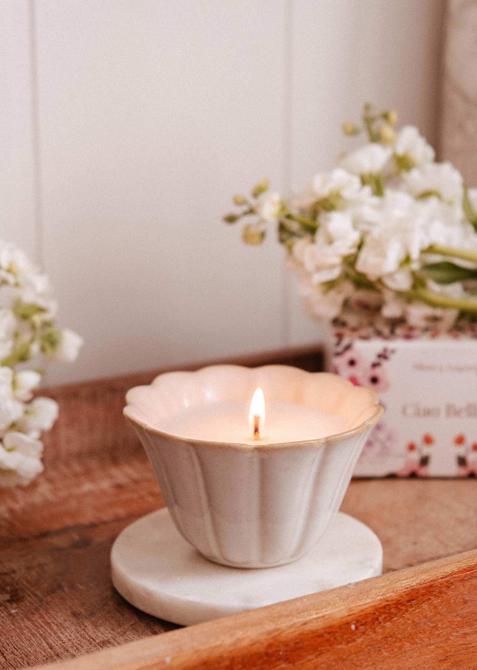The Ciao Bella - Reusable Candle by Mimi & August, in a white scalloped ceramic cup on a round marble coaster, is displayed with white flowers and a floral box in the background on a wooden tray.