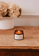 The Mimi & August Ciao Bella – 2 oz Soy Candle, hand-poured in Montreal, glows in a brown glass jar on wood beside a white vase with pale dried flowers.