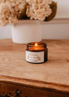 A lit Mimi & August Ciao Bella – 2 oz Soy Candle, hand-poured in Montreal, sits on a wooden surface with a blurred white vase of flowers in the background.