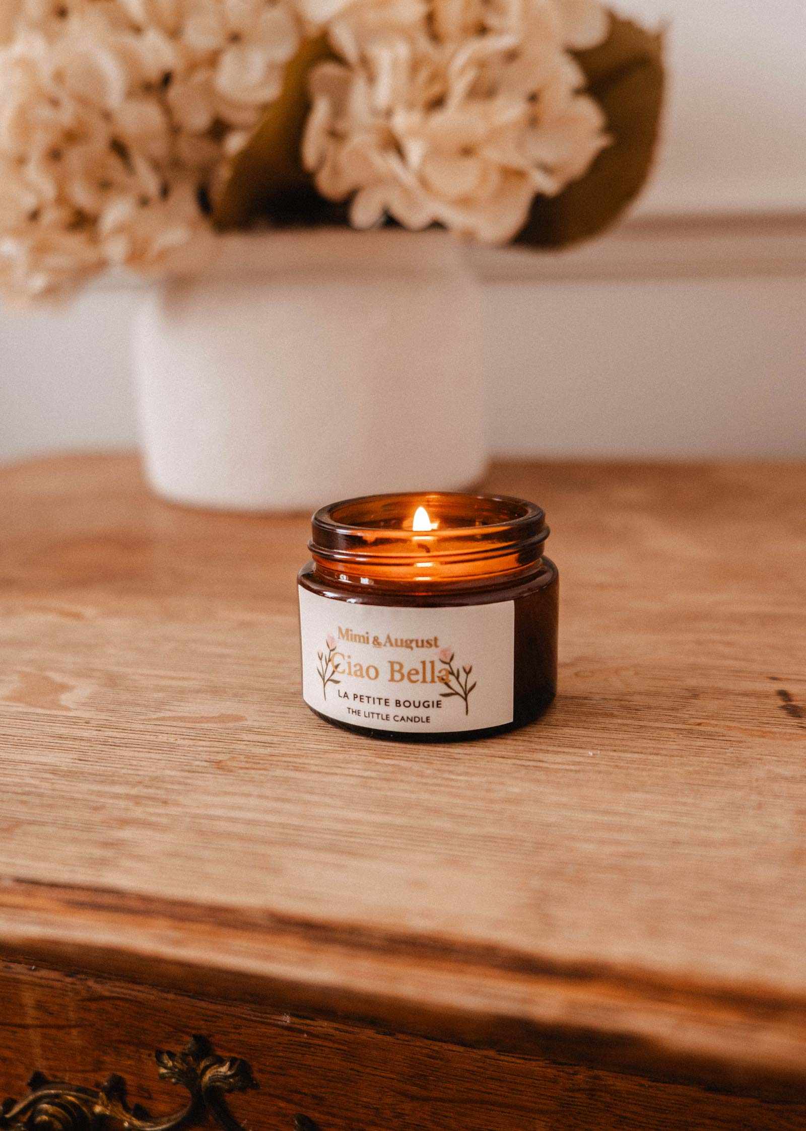A lit Mimi & August Ciao Bella – 2 oz Soy Candle, hand-poured in Montreal, sits on a wooden surface with a blurred white vase of flowers in the background.