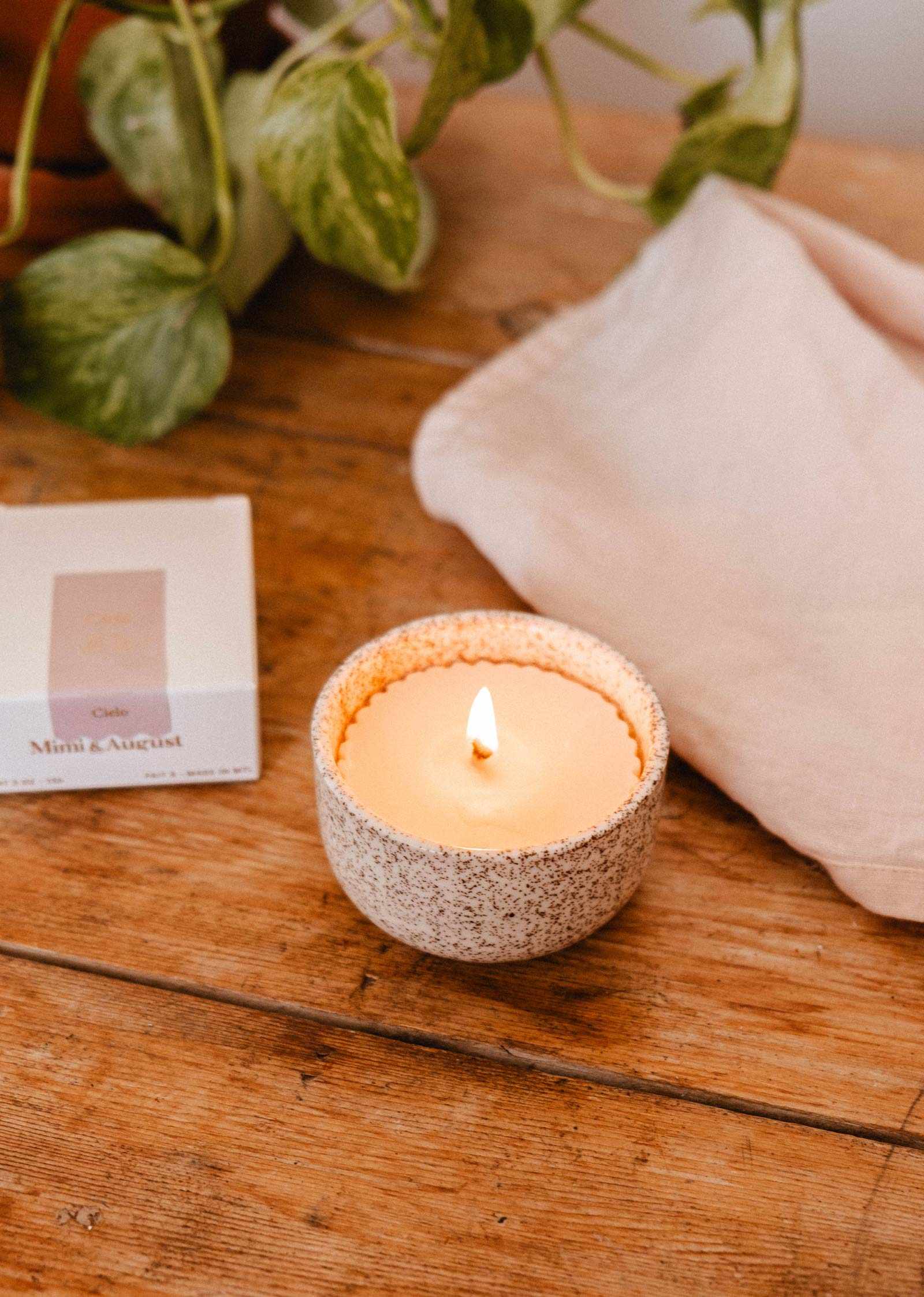 A lit Candle Refill Cielo by Mimi & August glows in a speckled ceramic holder on wood beside a white cloth and a box labeled "Mini August," with green leaves in the background.