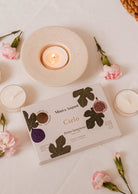 A Cielo Tealight Candle Set from Mimi & August, accompanied by pink flowers and flickering flames, creates a cozy ambience with its warm scent on a white table.