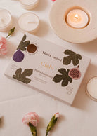 The Cielo Tealight Candle Set by Mimi & August, when paired with soft pink flowers and shimmering tea lights, creates a cozy atmosphere. Its warm, comforting scent enhances the inviting ambiance beside a lit candle on a saucer.