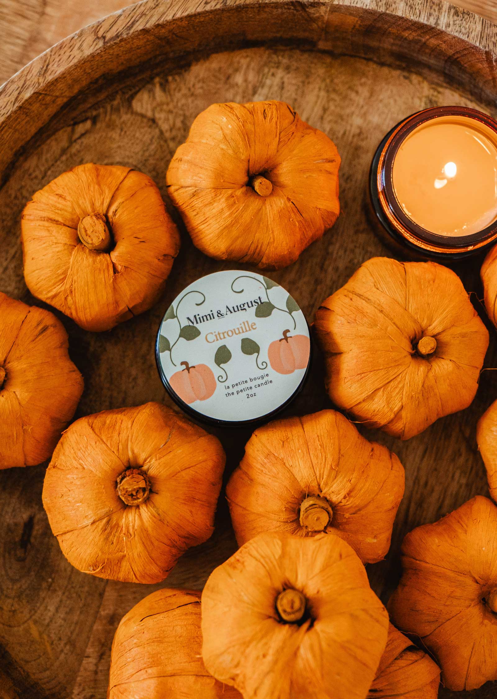 A collection of small orange pumpkins surrounds the Mimi & August Citrouille - Reusable Candle in a wooden bowl, exuding a warm and soothing pumpkin fragrance.