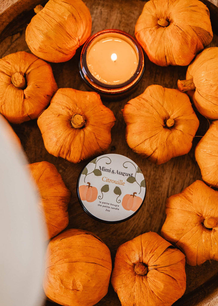 A wooden bowl featuring a lit Citrouille - Reusable Candle by Mimi & August, emitting a warm and soothing pumpkin fragrance, accompanied by faux orange pumpkins and a tin labeled "Mimi & August Citrouille.