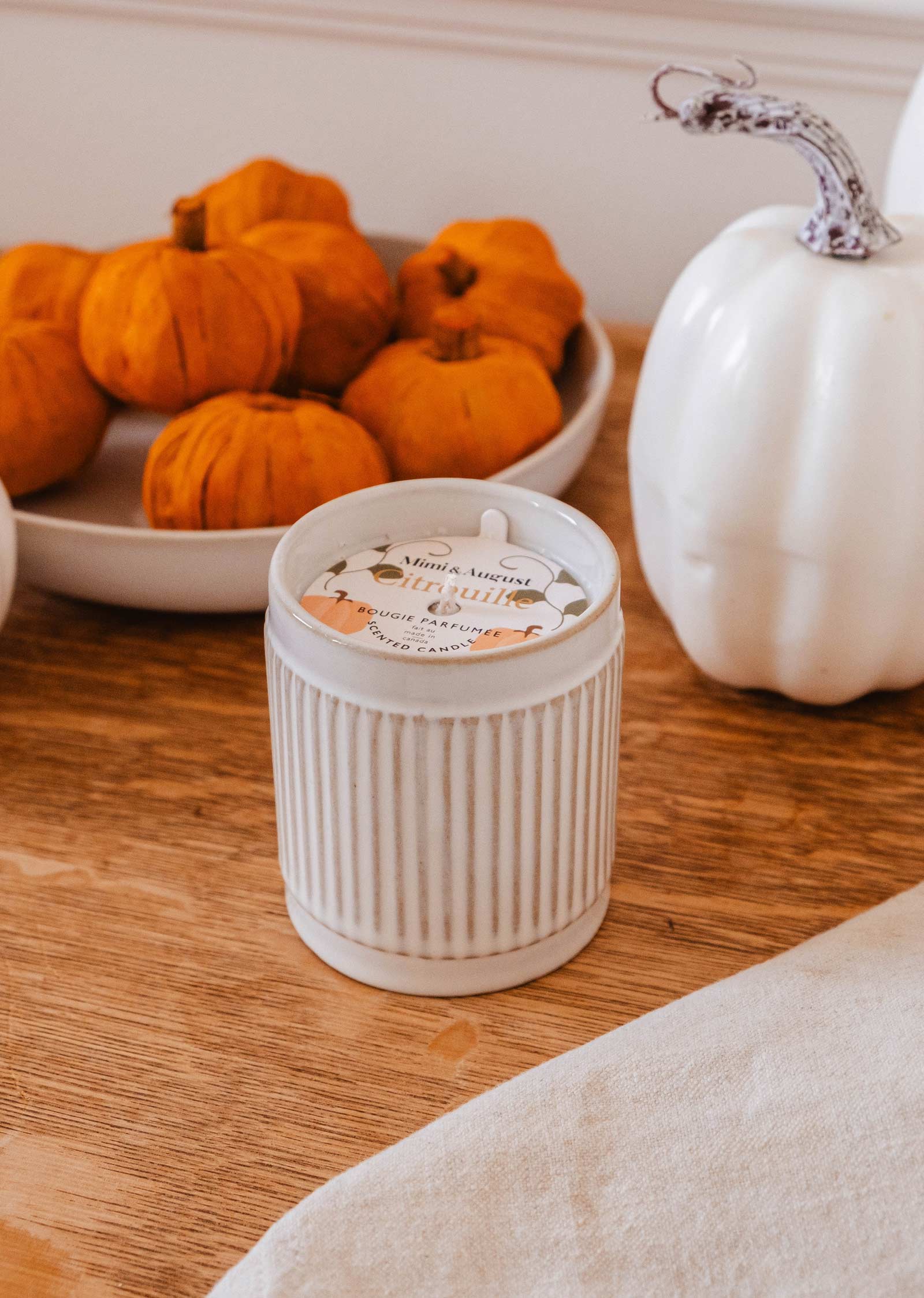 A white ribbed candle holder sits on a wooden surface, surrounded by small orange pumpkins in a bowl and a white pumpkin in the background. The Citrouille - Reusable Candle by Mimi & August emits a warm and soothing Pumpkin fragrance, enhancing the cozy autumn ambiance.