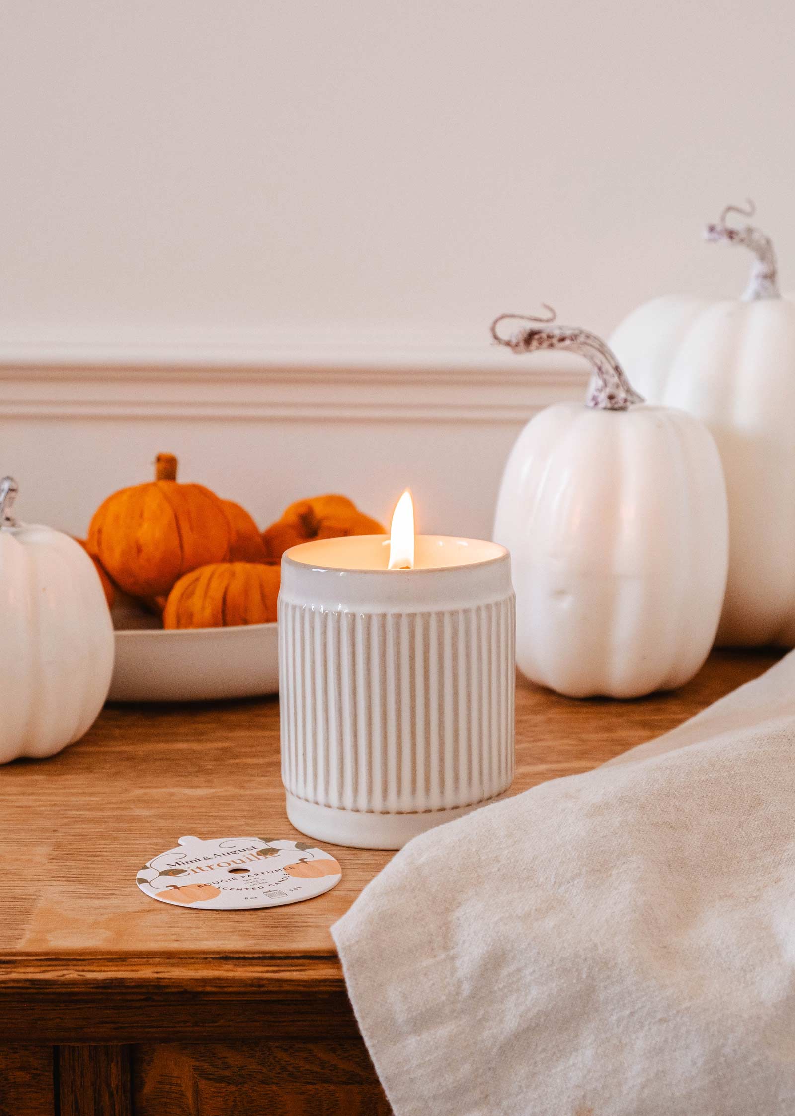 A lit Citrouille - Reusable Candle by Mimi & August is placed on a wooden table next to a cloth napkin, surrounded by white pumpkins and a bowl of small orange pumpkins, filling the air with a delightful pumpkin fragrance.