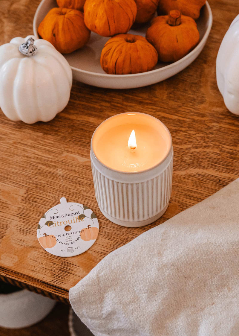 A lit **Citrouille - Reusable Candle** by **Mimi & August** sits on a wooden table next to a small dish of orange pumpkins and white pumpkin decorations. A card labeled "citrouille" is near the candle, emitting a delightful pumpkin fragrance.