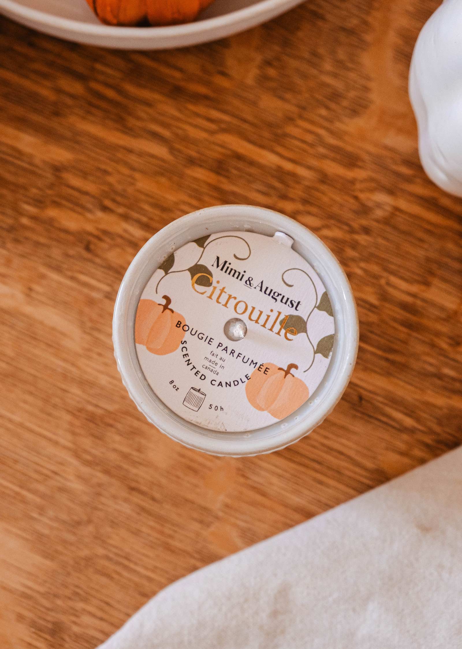 Top view of a scented candle in a tin, labeled "Citrouille - Reusable Candle" by "Mimi & August," placed on a wooden surface. The label, adorned with an illustration of pumpkins, promises a warm and soothing pumpkin fragrance. This reusable candle is perfect for cozy fall evenings.