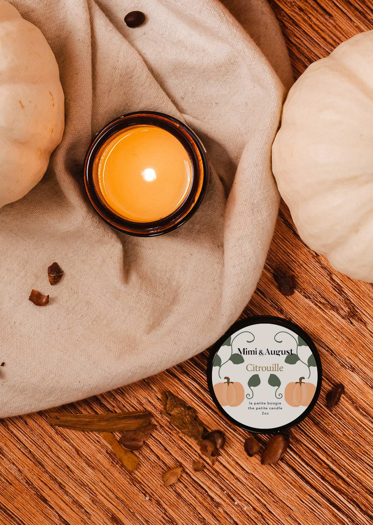 A Pumpkin candle 2 oz by mimi and august with pumpkins and spices on a table.