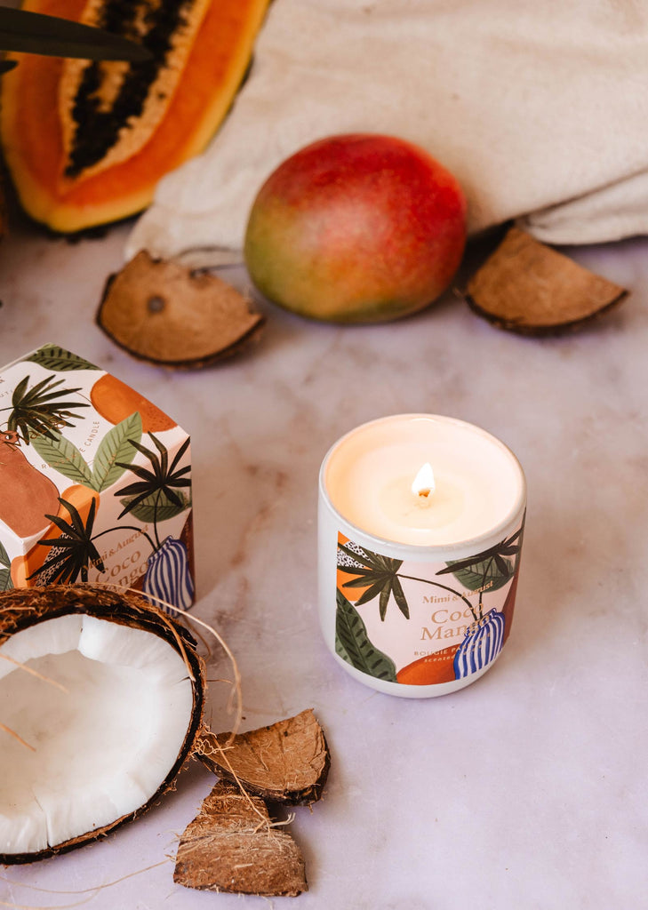 Une bougie réutilisable Coco Mango de Mimi & August dans un contenant décoré est posée sur une surface entourée de coquilles de noix de coco, d'une mangue, d'une tranche de papaye et d'une boîte aux motifs tropicaux, imprégnant l'air de son parfum tropical.