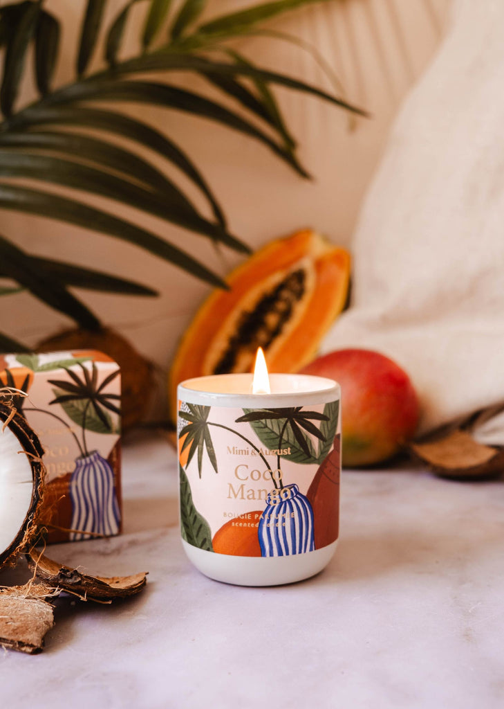 A lit tropical candle from Mimi & August labeled "Coco Mango - Reusable Candle" sits on a surface with a sliced papaya, a mango, and lush tropical leaves in the background, emitting an irresistible Coco Mango scent.